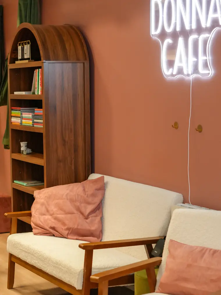 Le salon du café : un canapé blanc aux coussins rose poudré, une bibliothèque en bois et l’enseigne néon « Donna Café »